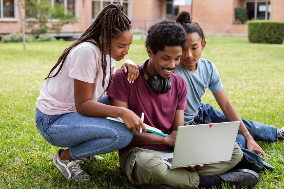 Três estudantes no jardim da universidade. Um aluno segura o notebook enquanto procuram o telefone FIES 2025