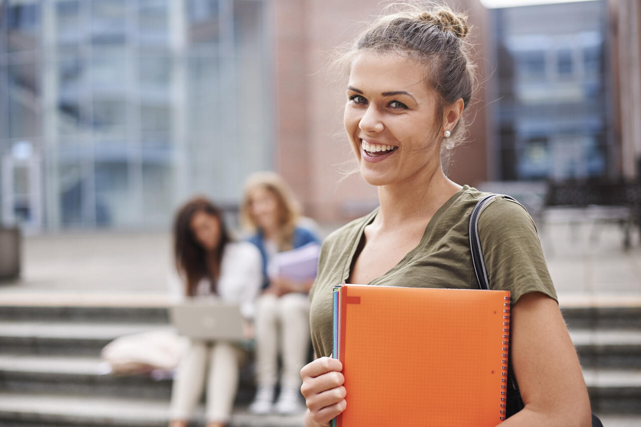 Estudante posa sorridente segurando cadernos no campus da universidade com colegas ao fundo 