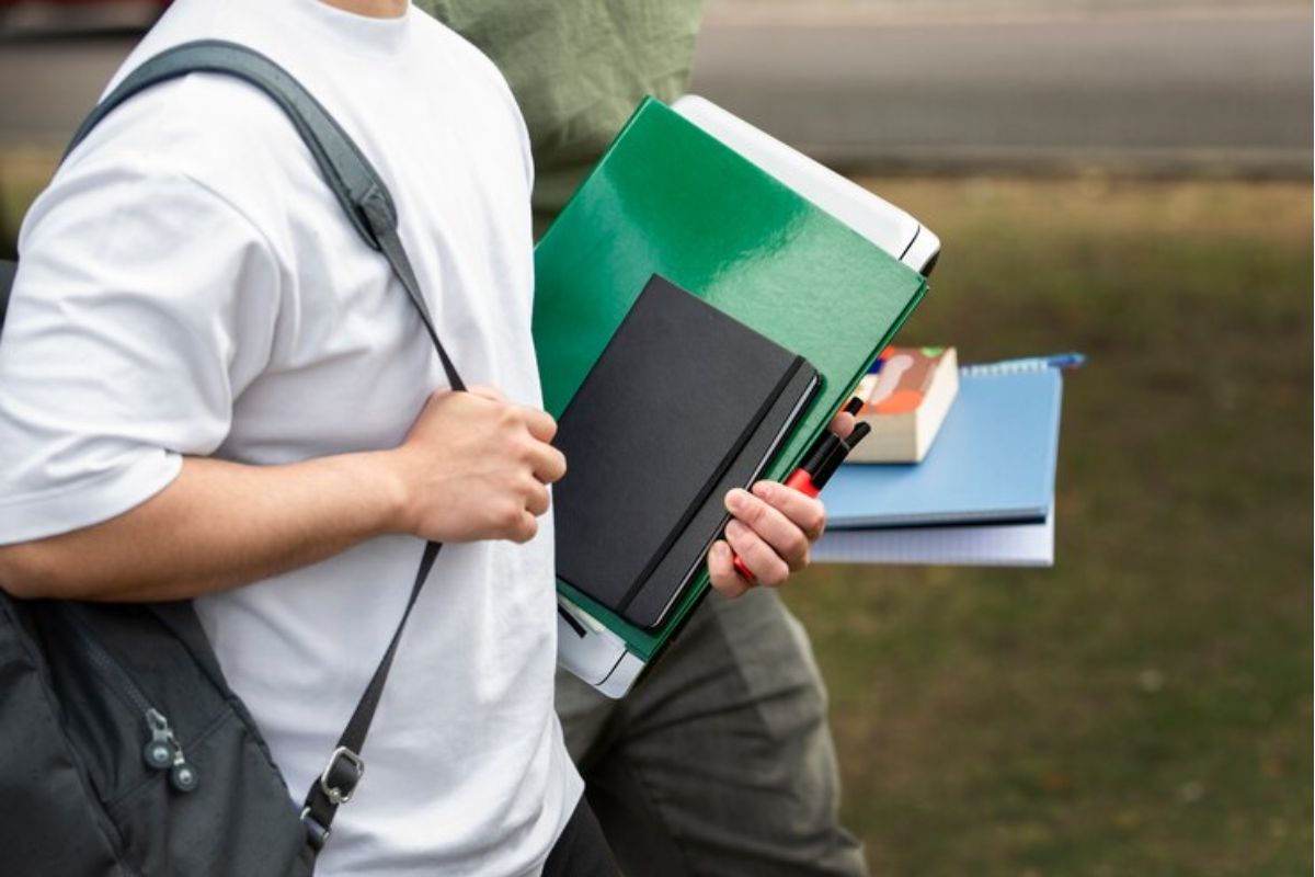 Dois estudantes caminham juntos com o material em mãos. Eles conversam sobre como renda familiar pode influenciar para você conseguir ou não o FIES