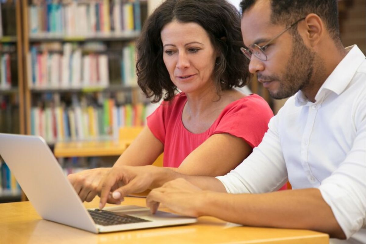 Dois estudantes em uma biblioteca consultam as informações sobre o Desenrola FIES 2025 em um notebook que está na mesa