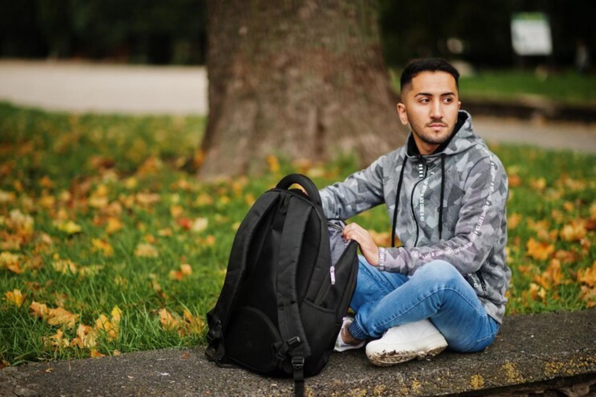 Um estudante sentado em um banco na praça com a mochila em sua frente