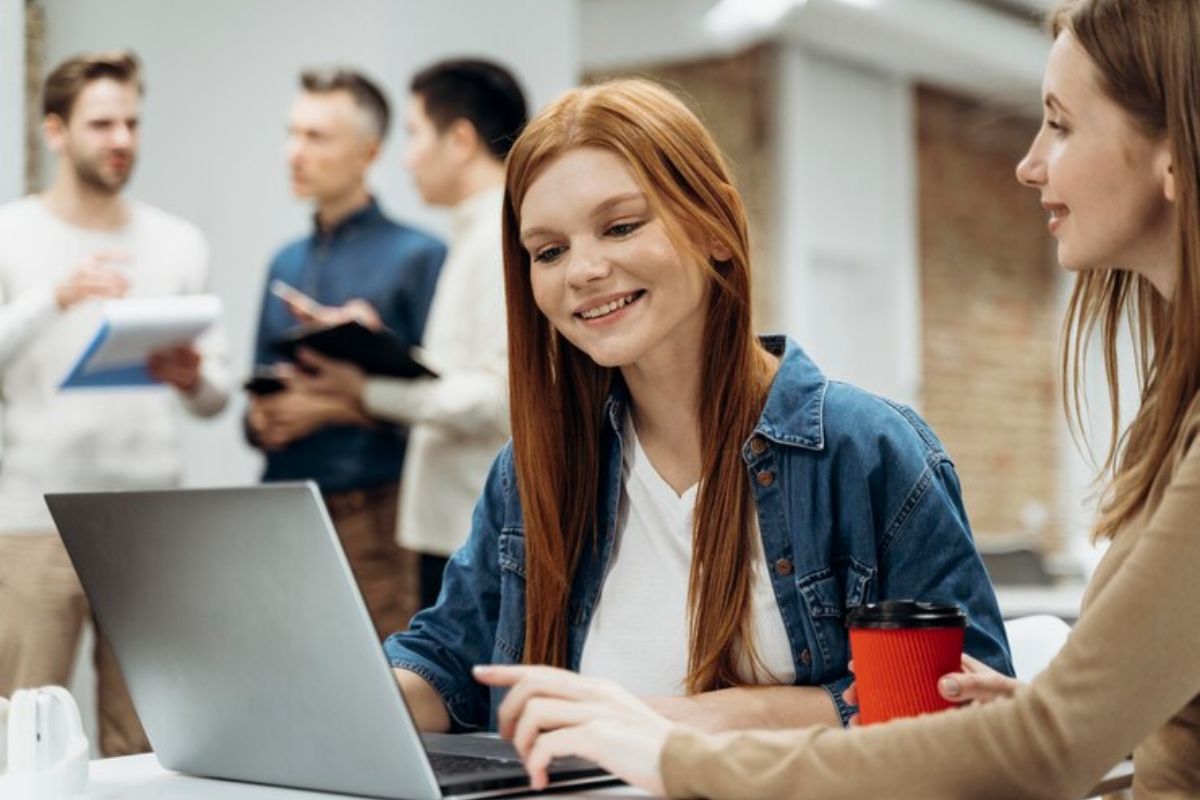 Uma estudante e sua amiga conversam em frente ao notebook. Ao fundo é possível ver dois estudantes ao fundo