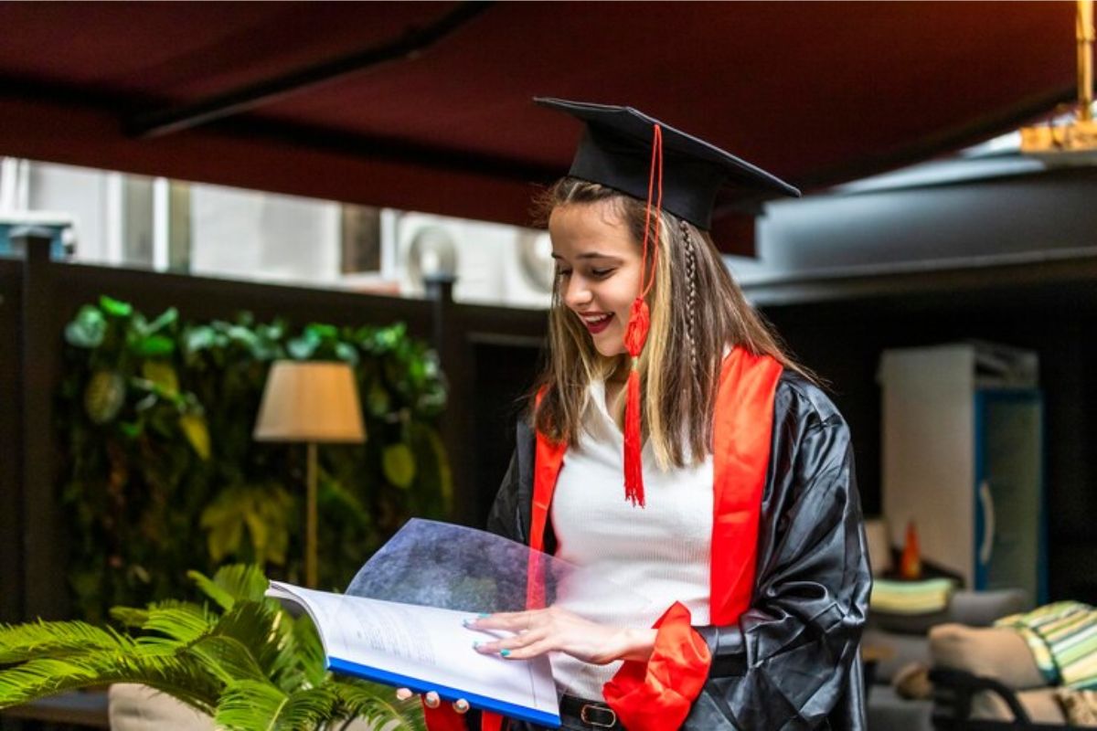 Uma estudante com roupa de formando olha para um caderno aberto que está em sua mão