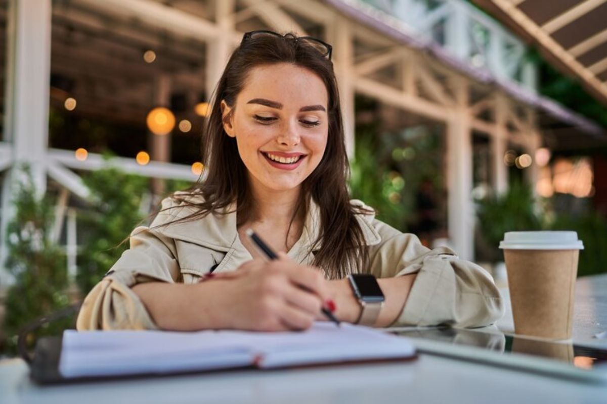 Uma estudante sentada na mesa fazendo anotações em seu caderno. Ao lado há um copo de café