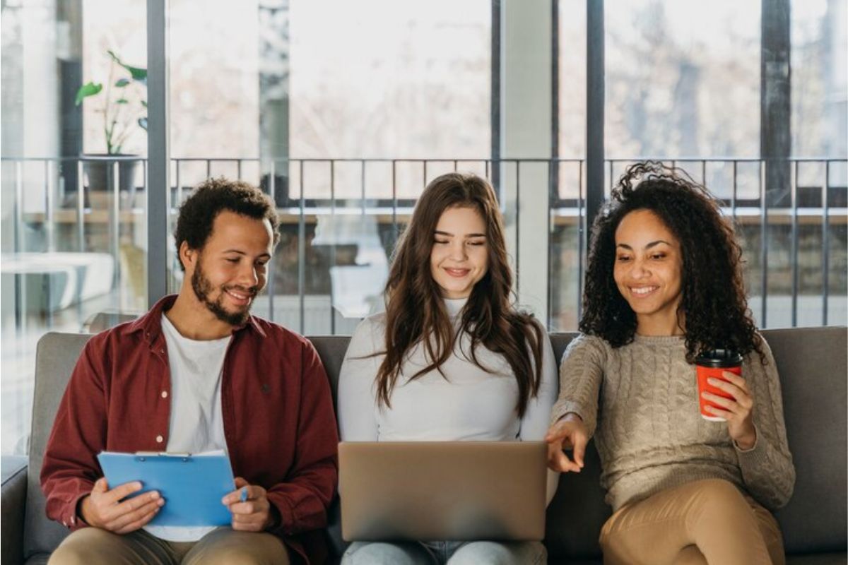 Três estudantes estão em um sofá. Uma amiga segura o notebook no meio dos dois e eles verificam a lista de espera do FIES 2025