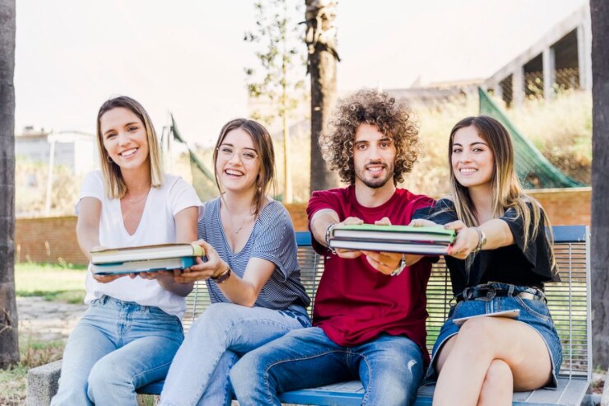 Três amigas e um colega estão sentados na mureta da universidade e segura cadernos enquanto sorriem para a foto