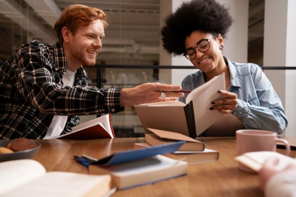 Dois estudantes fazem o trabalho juntos na biblioteca. A colega está com o livro aberto enquanto seu parceiro faz um apontamento na página