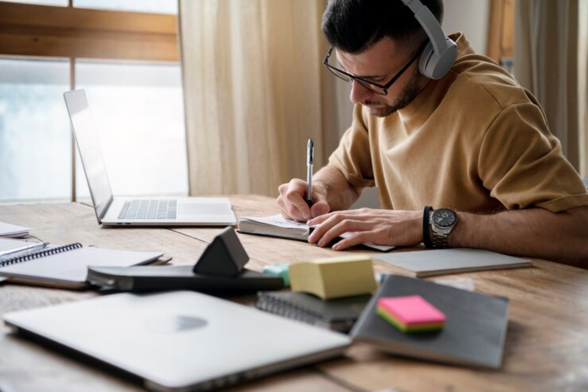 Um estudante em frente ao seu notebook no quarto. Ele faz anotações no caderno para ver como renegociar dívidas do FIES