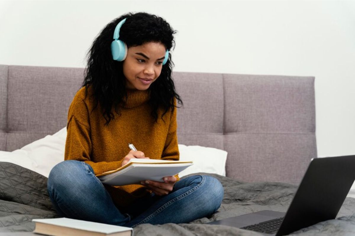 Uma estudante que está devendo o FIES usa o caderno para fazer anotações. Ela está sentada no sofá olhando para o notebook com um headphone 